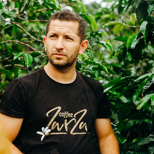 Man holding a red bucket amidst coffee plants wearing a black t-shirt with 'coffee Zara' branding.