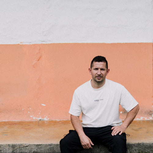 A man wearing a white crewneck t-shirt and black pants sits on a concrete ledge in front of a two-toned wall with white on top and orange on the bottom, next to a dark doorway.